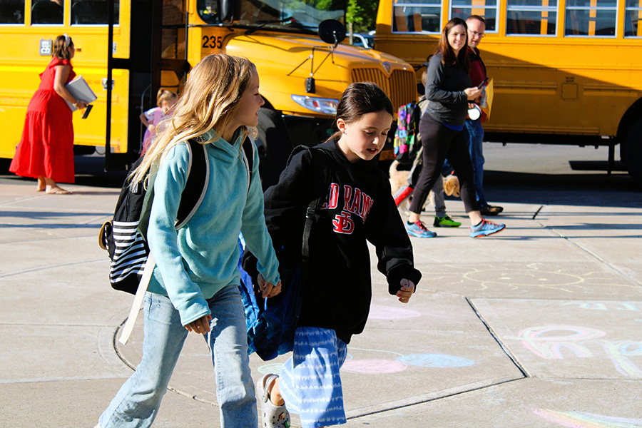 two students walk into Jamesville Elementary School wearing backpacks with buses in the background.