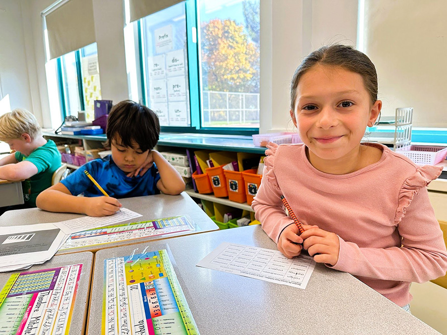 student smiles at camera while doing work at a desk.