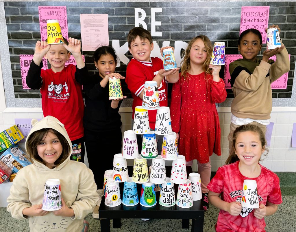 Moses DeWitt students stand next to decorated cup pyramid and smile while holding up coffee cups.