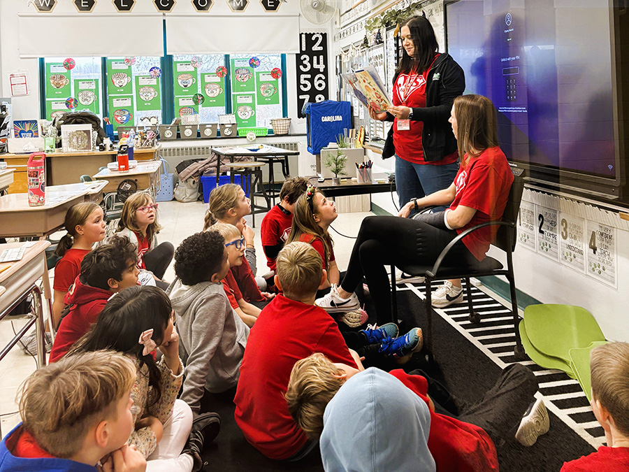 Students sit on classroom rug listening to teacher read a story.